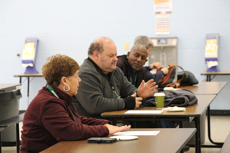 A group of three adults sit at tables with paper in front of them. They are talking.