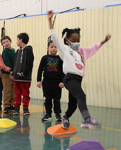 A group of younger elementary students stand in line and watch as a little girl steps from one brightly colored stepping stone to another. 