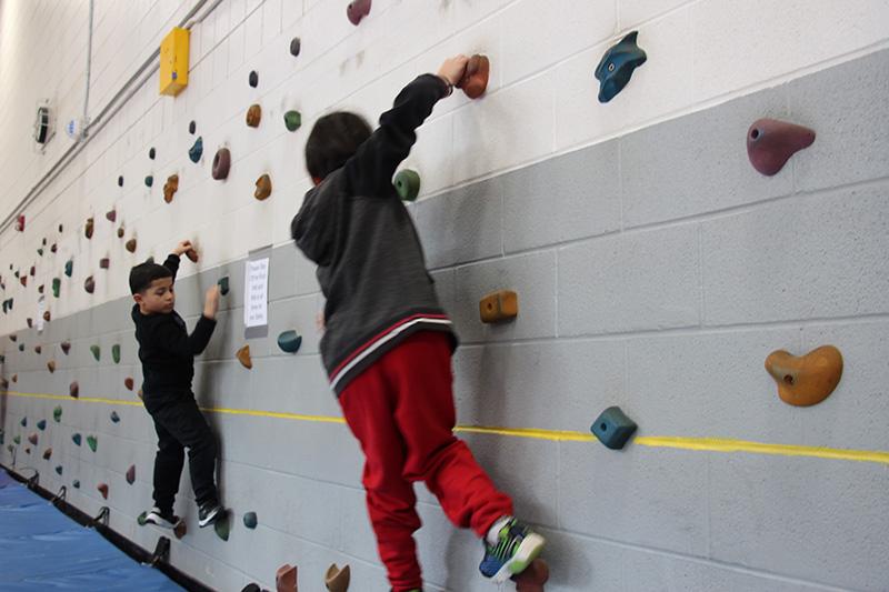 Young elementary students, dressed in sweat pants and sweatshirts, go sideways on a rock climbing wall. The hand and foot holds are different colors.