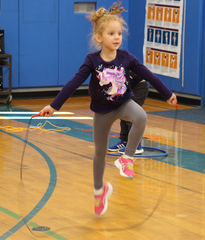A young elementary age girl, with her hair pulled up, wearing a blue long-sleeved shirt and gray pants and pink sneakers, jumps rope.