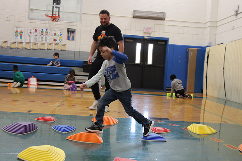 A younger elementary age boy, wearing a gray and blue long-sleeve shirt and dark pants, jumps from one brightly colored stepping stone to another. A man in a dark shirt stands behind watching him.