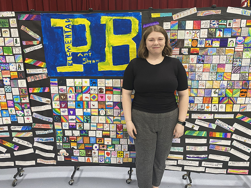 A woman stands in front of a large board of artwork.