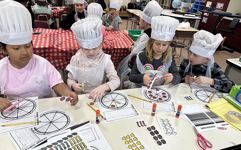 A group of four third-grade students wearing aprons and white chef hats work on fractions on paper that looks like a pizza.