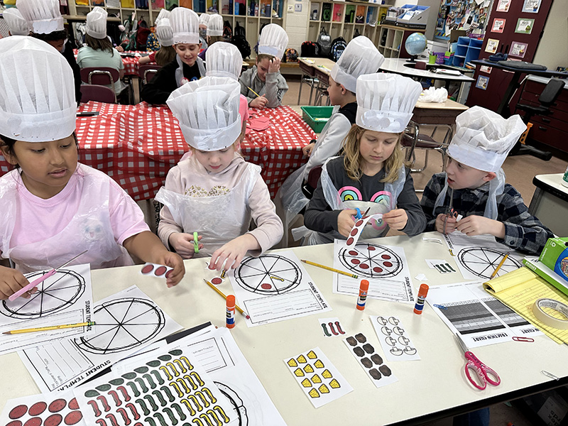 A group of four third-grade students wearing aprons and white chef hats work on fractions on paper that looks like a pizza.
