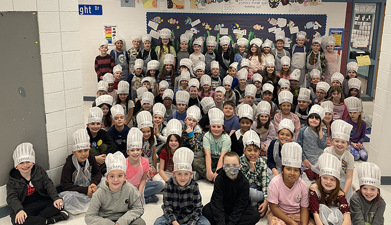 A group of about 80 third-grade students, all wearing white chef hats, sit and stand in a hallway.