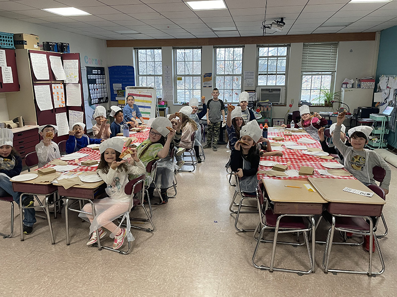 Long lunch tables filled with third grade kids eating pizza. There are red and white checked table clothes on the tables.