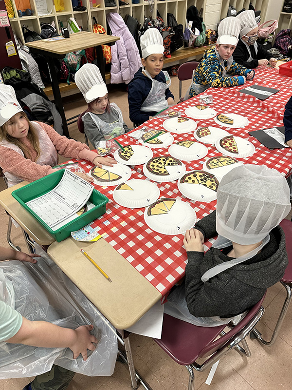 A group of five third-grade kids, all wearing white chef hats, working on fractions in the shape of pizza. There is a red and white check table cloth on the table.