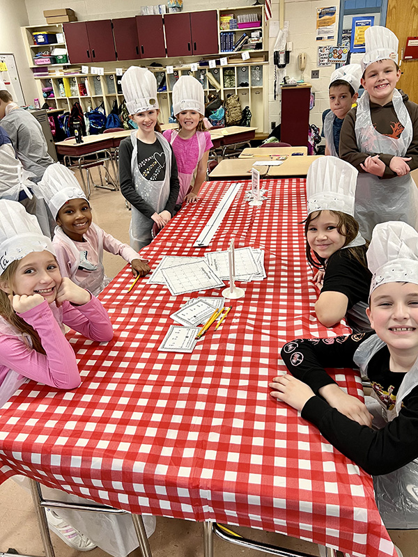 Six third-grade students sit at a table with a red and white check table cloth. They are all wearing a white chef's hat.