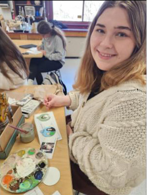 A high school student with blonde hair wearing a tan sweater paints an ornament.