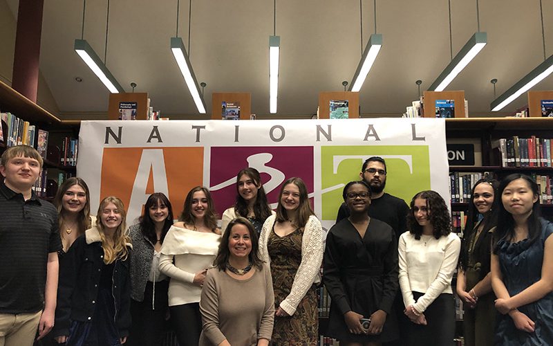 A group of 12 high school students stand in front of a large banner that says National Art Honor Society. There is a woman in front.