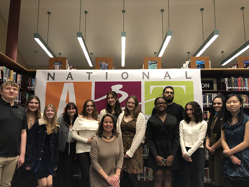 A group of 12 high school students stand in front of a large banner that says National Art Honor Society. There is a woman in front.