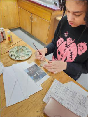 A high school student in a black shirt with a pink elephant on front, paints an ornament.