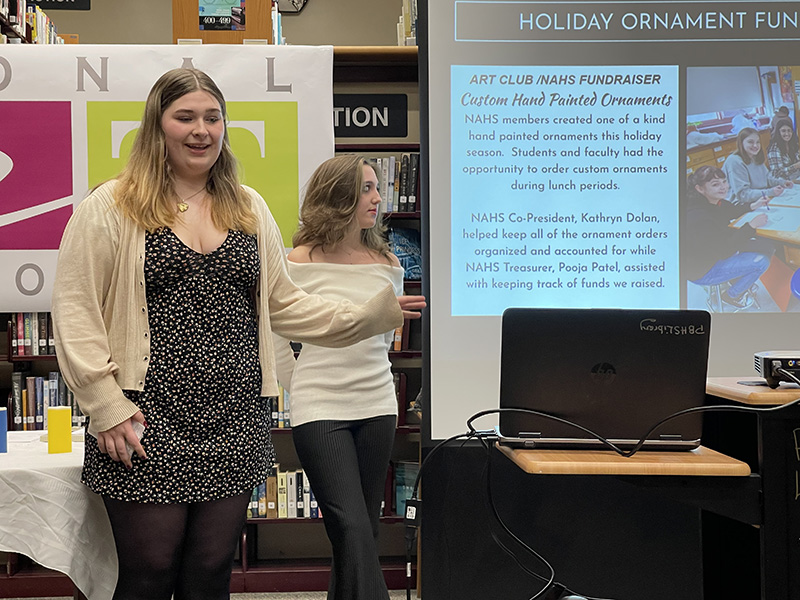 Two high school girls stand in front of a slide show presenting it.