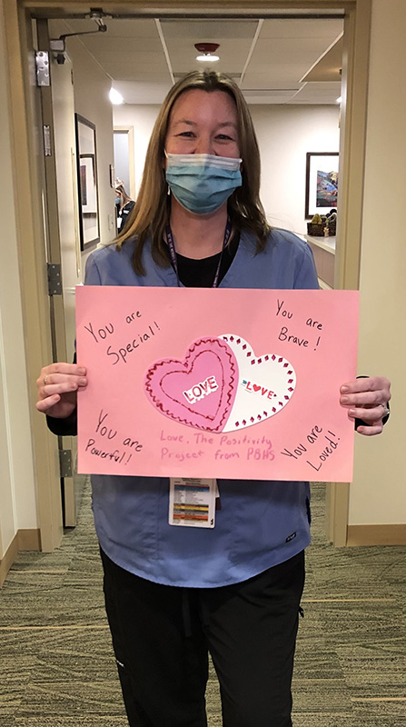 A woman in blue scrubs holds a large pink valentine with pine and white hearts on it.