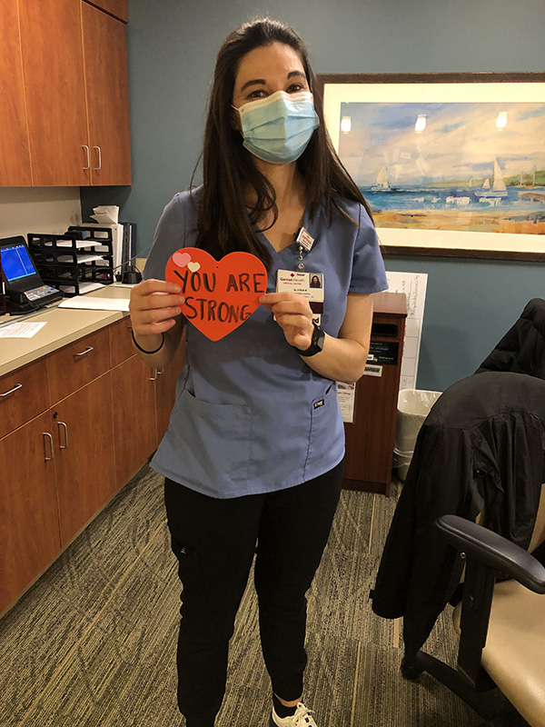 A woman in scrubs holds up a red heart that says You Are Strong.