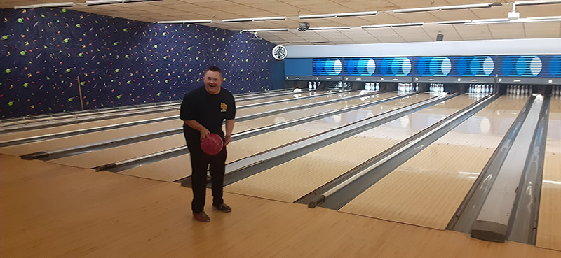 A young man wearing dark colored clothes is laughing, while holding an orange bowling ball. He is standing at the edge of a bowling lane.