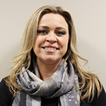 A woman smiles. She has long blonde hair and a gray and white scarf around her.