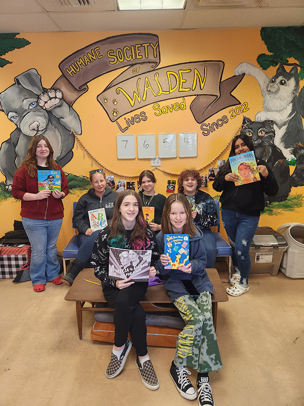 A group of six middle school students, four sitting on chairs and two standing all holding books, are with one adult, also sitting holding a book. Behind them is a wall painted with a puppy and a kitten. It says Humane Society of Walden Lives Saved Since 2012 7654.