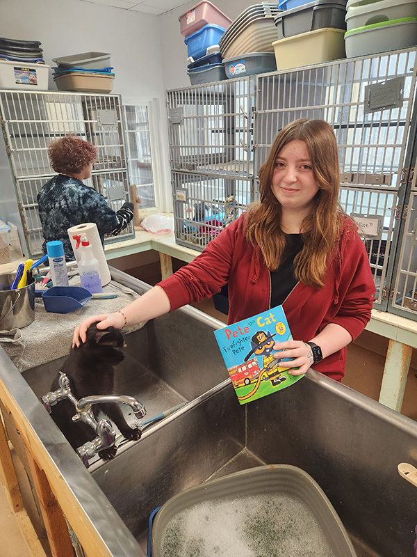 A middle school student holds a book that says Pete the Cat while petting a black cat in a large sink. The  student is smiling. She has long brown hair and is wearing a black shirt and red sweatshirt.