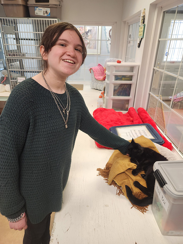 A middle school student  with dark hair pulled back, wearing several necklaces and a black shirt, smiles as she pets a black cat.