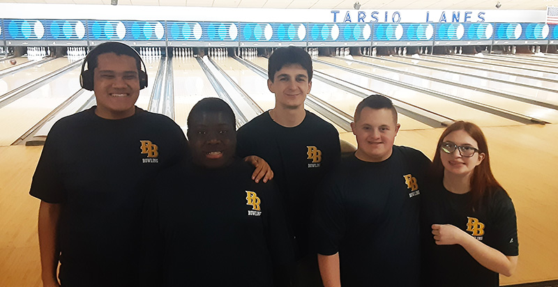 A group of five - four young men and one young woman - stand together with bowling lanes in the background. They are smiling.