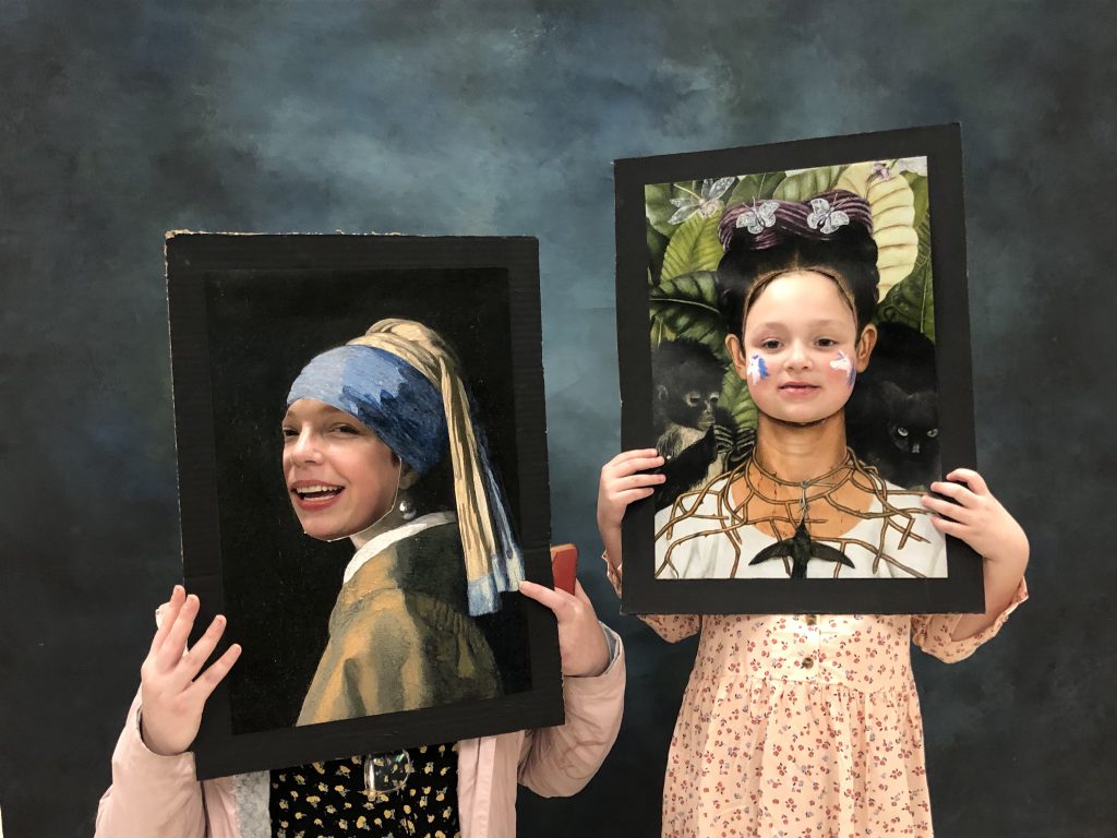 Two elementary students hold up copies of classic pieces of art with their faces on them.