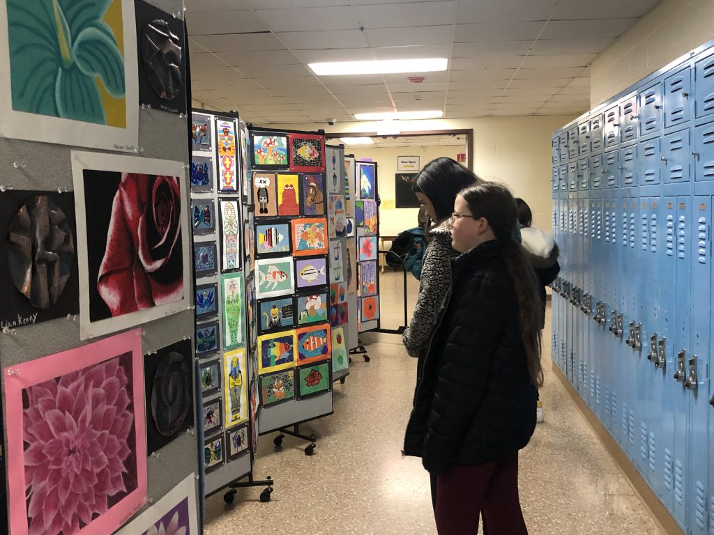 Adults walk down a hall looking at pieces of art on walls.