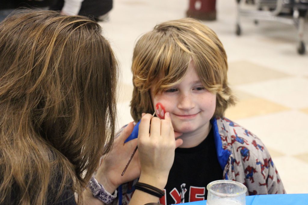 An elementary boy sits and gets his face painted.
