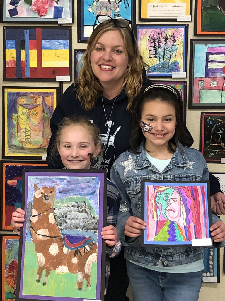 Two elementary girls hold paintings they made and smile. There is a woman behind them also smiling. The wall behind them is filled with paintings.