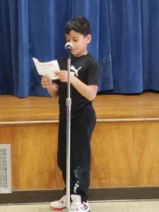 An elementary boy dressed in black shirt and sweat pants  reads from a piece of paper at a microphone.