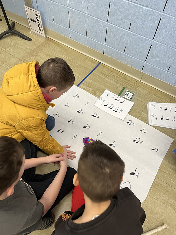 Three fourth-grade students hover over a large piece of paper with musical notes on it with a computer mouse. The view is from above.