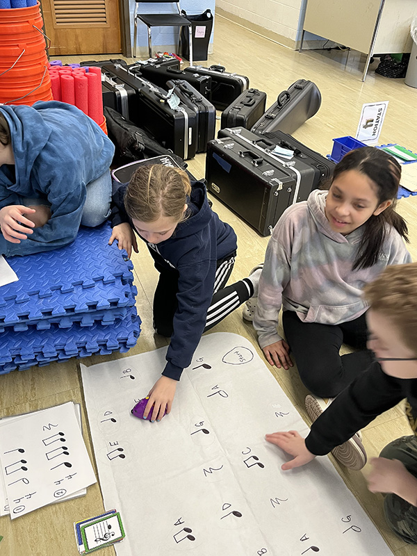 Four fourth-grade students hover over a large piece of paper with musical notes on it with a computer mouse.