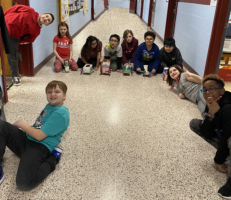 A group of 10 fourth-grade students in a school hallway with the race cars they created from recycled materials. all are smiling. Six are in the center with their cars ready to race them.