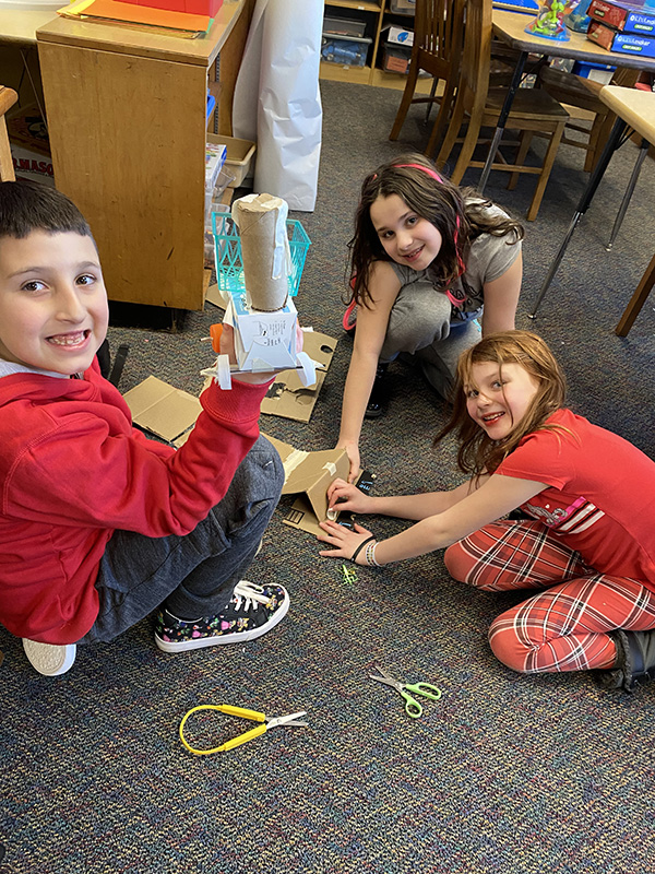 Three fourth-grade students are on the floor making their cars from recycled materials.