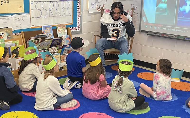 A high school student in his football uniform sits in front of a class of young elementary students reading to them. The students are sitting on a colorful rug wearing paper hats listening.