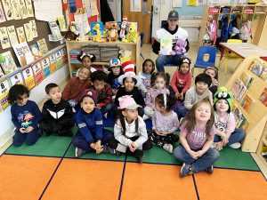 A class full of young elementary students  smiles and a high school kid in his football jersey sits with them . 