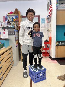 A high school student wearing a white football jersey that says Pine Bush with the number 4 on it smiles broadly as a young elementary student stands next to him, standing on a large box. Both are smiling.