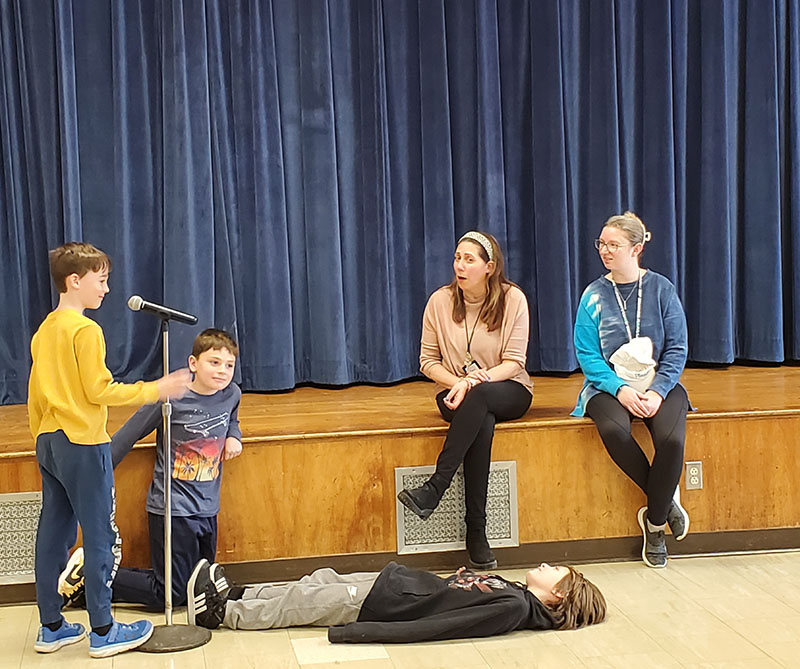 Two adults sit to the right on a stage. Three elementary age boys are performing a skit in front of them. One boy is laying on the floor and the other two are at a microphone.