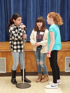 Three older elementary age girls stand in front of a stage at a microphone.