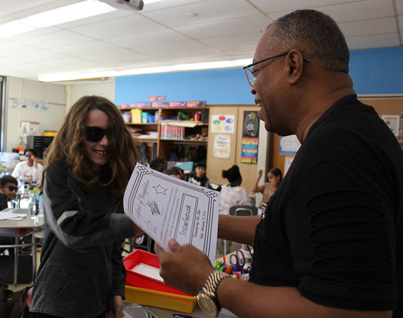 A man with glasses and wearing a dark shortsleeve shirt, hands a certificate to a fifth-grade boy with long blonde hair and wearing dark sunglasses.