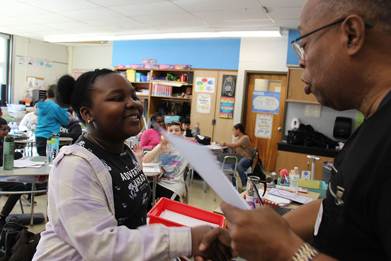 A man with glasses and very short hair hands a certificate to a girl who is smiling broadly. She has pig tails and is wearing a dark shirt with a pink sweatshirt over it.