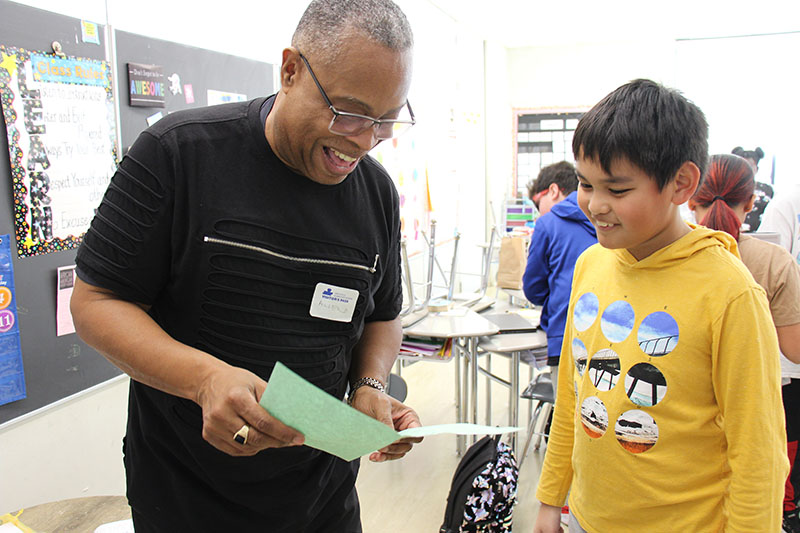 A man with a dark shirt and short hair and glasses smiles as he looks over a green sheet of paper with a fifth-grade boy wearing a yellow shirt. He is smiling too.