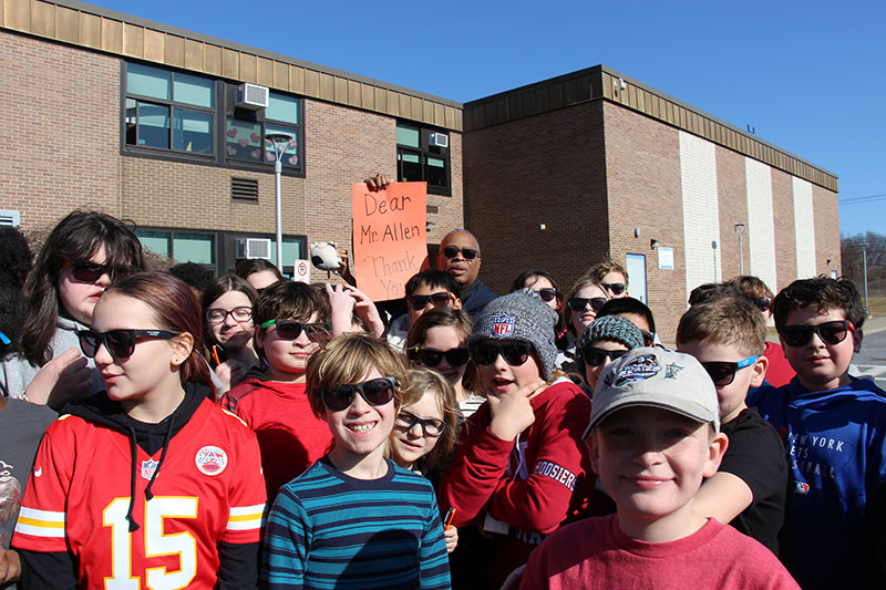 A large group of fifth-grade kids, mostly boys huddled around a man who is holding up a large orange card that says Thank you Mr. Allen.