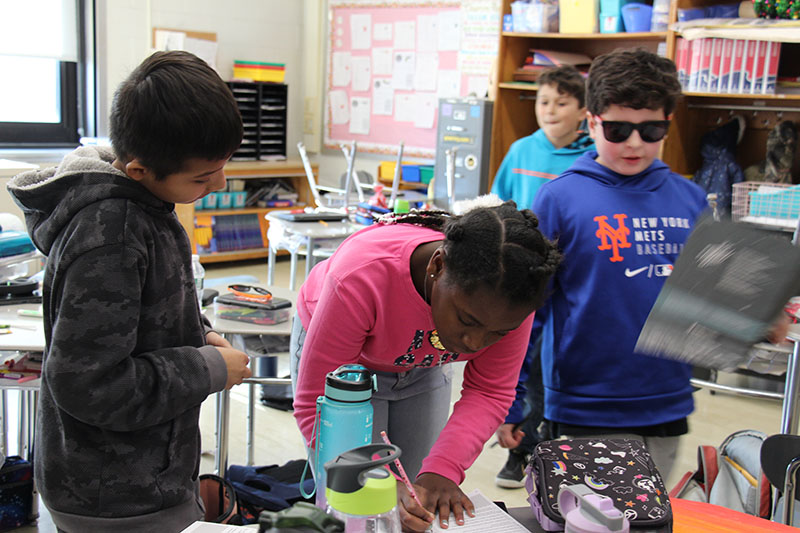 Three fifth-grade students work together in their classroom. A girl in a pink shirt is bending over the desk writing, while two other kids look on.