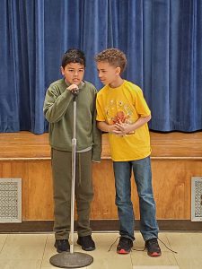 Two elementary age boys stand at a microphone and talk.