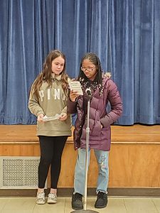 Two elementary girls stand at a microphone and talk.