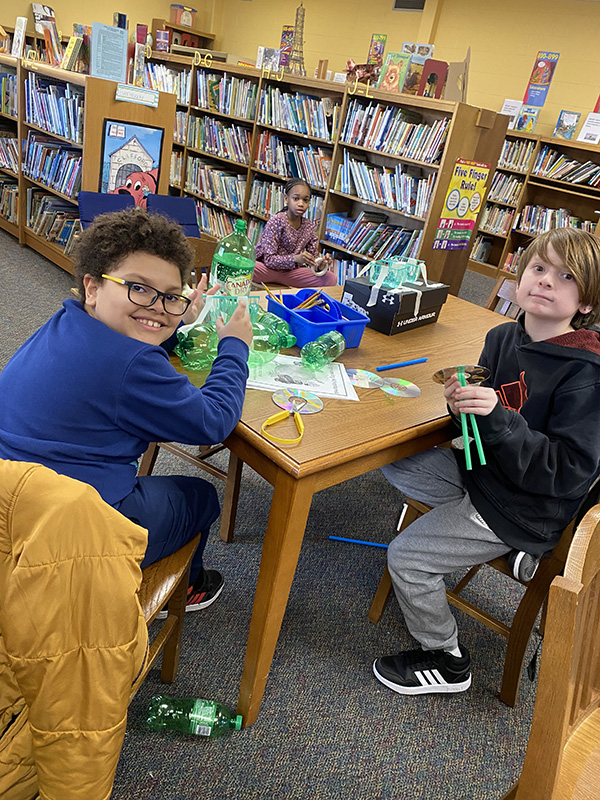 Three fourth-grade kids sit at a table with lots of recycled materials on it.