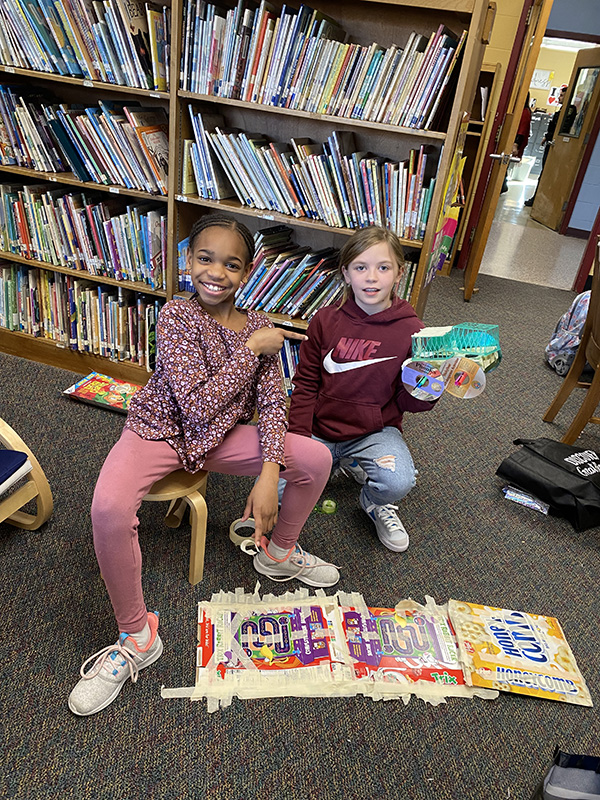 Two fourth-grade kids sit together working on their race cars made from recycled materials.