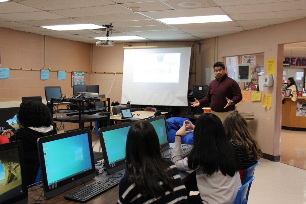 A man stands at the front of a classroom at a projection screen. The students are sitting at tables with rows of computer screens.