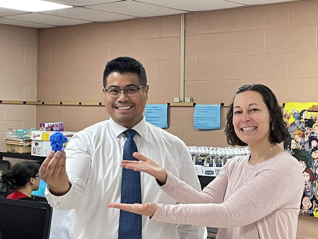 A man with dark hair and glasses, wearing a white shirt and blue tie smiles and holds up a blue spike protein that was 3D printed. A woman with chin-length dark hair smiles and points to the structure.